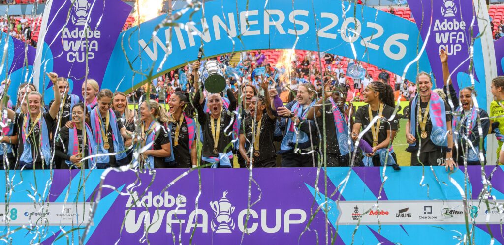 A women's team lifting the Women's FA Cup trophy amid confetti at Wembley.